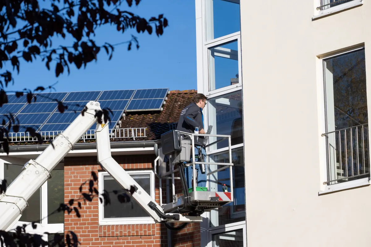 Ein Mitarbeiter von G&W reinigt von der Hebebühne aus ein Fenster, dahinter ein Dach mit Solaranlage unter blauem Himmel.