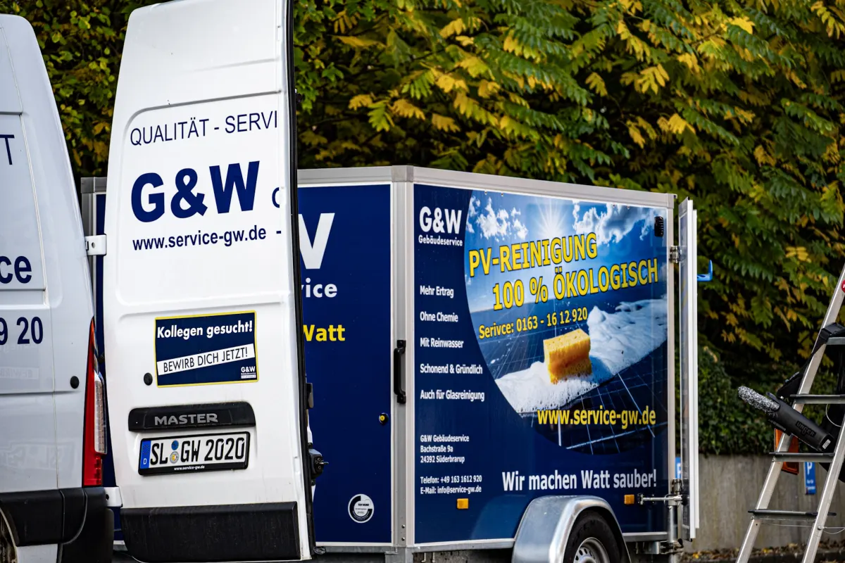Ein weißer G&W-Transporter mit Anhänger, daneben eine Leiter, vor herbstlichen Bäumen.